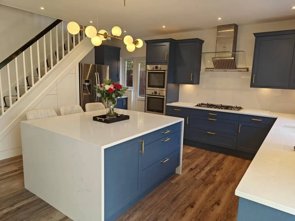 Navy blue shaker kitchen island with white quartz worktop and brass handles beneath open staircase, Bromley